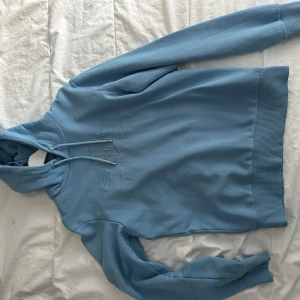 Blå hoodie från GANT - Säljer en snygg blå hoodie från GANT med broderad logga på bröstet. Tröjan har långa ärmar och en klassisk känguruficka framtill. Perfekt för en avslappnad stil.