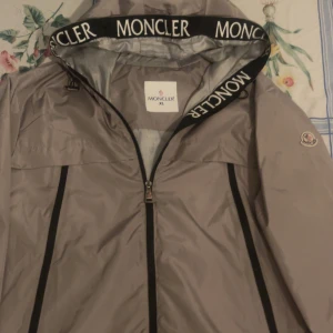 Grå vindjacka från Moncler - Snygg grå vindjacka från Moncler med dragkedja och huva. Jackan har Moncler-logga på ärmen och svarta detaljer längs dragkedjorna. Den är storlek XL men i svenskt så är de L. Perfekt för blåsiga dagar och stilrena outfits.