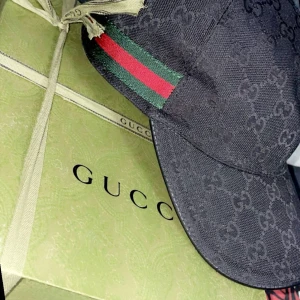 Svart keps från Gucci - Helt ny, Snygg svart keps från Gucci med det klassiska GG-mönstret. Kepsen har en grön och röd randdetalj på sidan. Perfekt för att ge din outfit en lyxig touch.