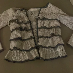 Vit stickadkofta från Odd Molly - Säljer en vacker ljus beige stickadkofta från Odd Molly med volanger och fina detaljer. Koftan har långa ärmar och en liten rosett på baksidan. Perfekt för att ge en romantisk touch till din outfit.