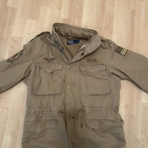 Beige jacka från Ralph Lauren - Säljer en beige jacka från Ralph Lauren med broderade detaljer och flera fickor. Jackan har en militärinspirerad design med märken på ärmarna och bröstet. Perfekt för en cool och avslappnad stil. Nypris runt 5500 säljer den för 1399 använd 3 gånger storlek M