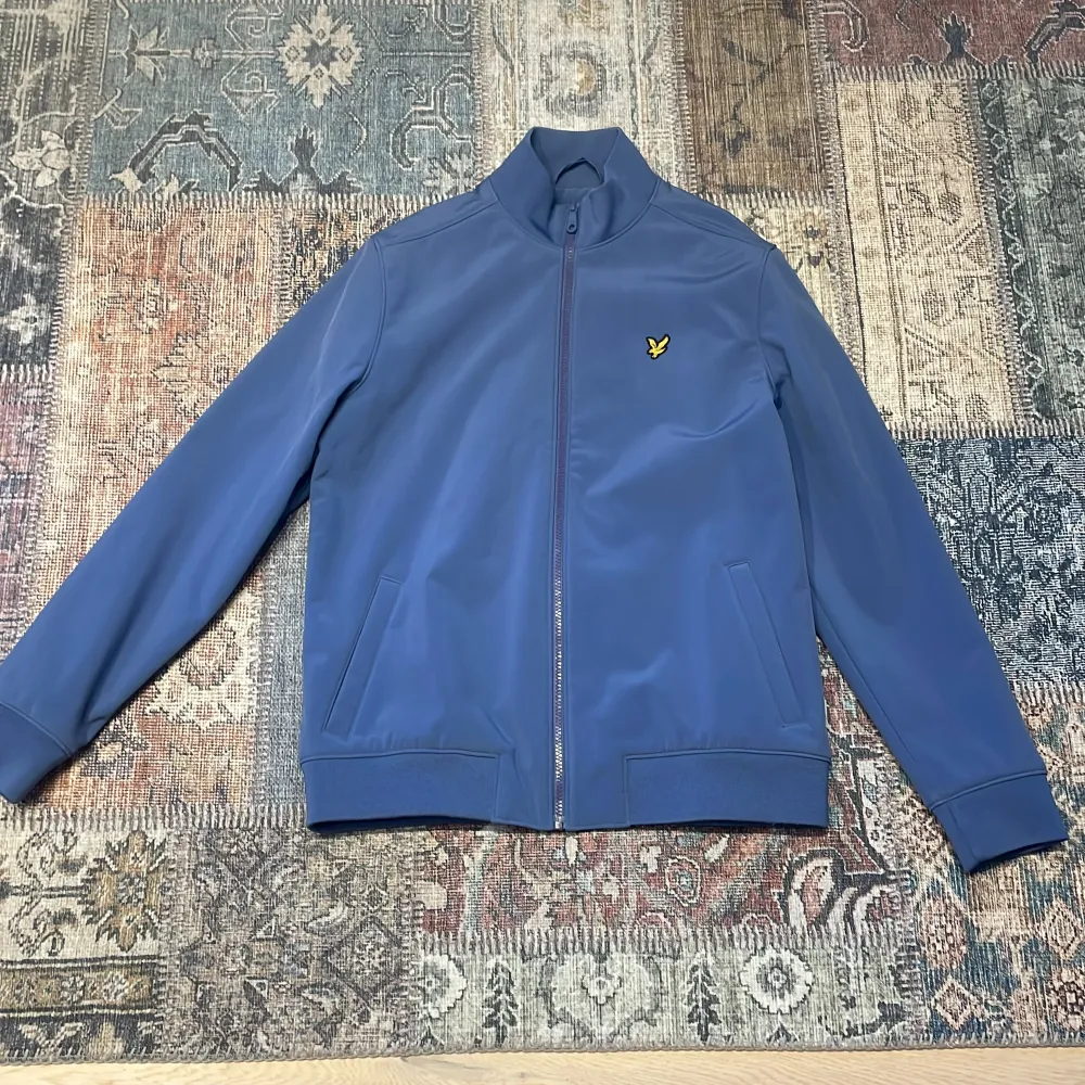 Säljer en stilren blå jacka från Lyle & Scott med dragkedja och ribbade detaljer. Jackan har en broderad logotyp på bröstet och långa ärmar. Perfekt för en casual look.. Takit.