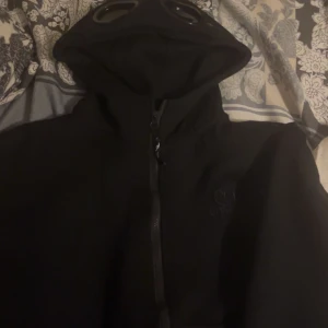 Svart jacka med huva från C.P. Company - Säljer en svart jacka från C.P. Company med unik design. Jackan har en huva med inbyggda glasögon och dragkedja framtill. Perfekt för en cool och annorlunda stil. Passar bra för höst och vår.