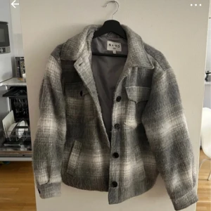 Grå rutig jacka från NA-KD - Säljer en stilren grå rutig jacka från NA-KD. Jackan har en klassisk krage och knappar framtill. Den är perfekt för kyligare dagar och har en mjuk och bekväm känsla. Passar bra till både jeans och kjol.