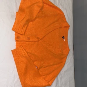 Orange stickad kofta - Säljer en härlig orange stickad kofta med knappar framtill. Koftan har långa ärmar och ribbade detaljer vid ärmslut och nederkant. Perfekt för att liva upp garderoben med en färgklick! 🍊