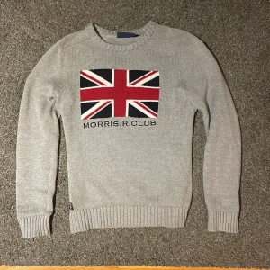 Grå stickad tröja från Morris  - Snygg grå stickad tröja från Morris  med ett flaggmotiv insytt på bröstet. Tröjan har långa ärmar och ribbade muddar vid ärmslut och nederkant. Nypris 1300kr. Säljer för 399. Inga defekter.