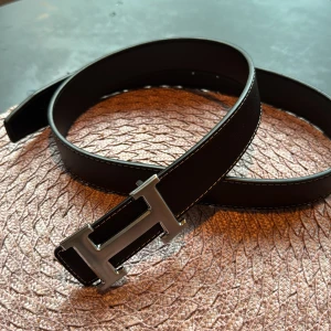 Svart bälte med silverfärgat spänne - Snyggt svart bälte i skinn med ett stilrent silverfärgat H-format spänne. Bältet har vita sömmar längs kanterna som ger en fin kontrast. Perfekt för att ge en elegant touch till din outfit.