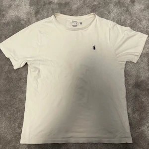 Vit t-shirt från Ralph Lauren - Säljer en klassisk vit t-shirt från Ralph Lauren med den ikoniska loggan broderad på bröstet. T-shirten är i bomull och har en normal passform med korta ärmar. Perfekt för en stilren och avslappnad look.