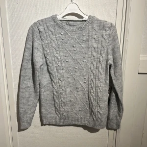 Grå stickad tröja från LC Waikiki - Säljer en snygg grå stickad tröja från LC Waikiki. Tröjan har ett klassiskt flätmönster och små dekorativa bollar på framsidan. Den är långärmad med ribbstickade muddar vid ärmslut och nederkant. Perfekt för kyliga dagar!