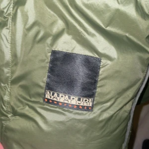 Grön pufferjacka från Napapijri - Säljer en grön pufferjacka från Napapijri. Jackan har en stilren design med märkets logga på bröstet. Perfekt för kyligare dagar med sin vadderade och quiltade stil. Den har en dragkedja framtill för enkel stängning. Skick 10/10