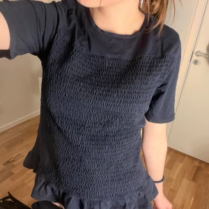 Mörkblå topp med volangdetalj - Säljer en mörkblå topp med korta ärmar och en snygg volangdetalj vid nederkanten. Toppen har en smockad textur framtill som ger en unik look. Perfekt för att piffa upp vardagsstilen!❤️