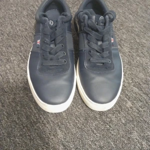 Mörkblå sneakers från Henri Lloyd - Snygga mörkblå sneakers från Henr lloydi med Union Jack-detalj på sidan. Skorna har en vit sula och klassisk snörning. Perfekta för en stilren look.
