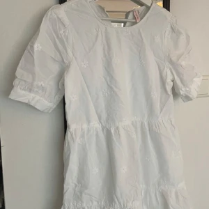 Studentklänning  - Charmig vit klänning med korta ärmar och ett blommigt mönster. Perfekt för studenten. Klänningen har en luftig passform och är tillverkad i ett lätt material, sparsamt använd.
