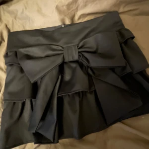  kjol med rosett  - Snygg svart kortkjol med en stor rosett där fram💋 Kjolen har en volangeffekt och en dragkedja baktill! Ser väldigt elegant ut och är i bra material, skriv gärna om ni har frågor eller prisförslag!!  Längd: 32 cm. Midja: 37 cm. 