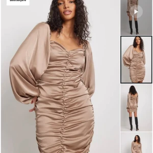 Beige kortklänning med ballongärmar - Säljer en superfin beige kortklänning med ballongärmar. Klänningen är i ett glansigt material som ser ut som satin och har en tight passform. Den har en snygg rynkad detalj framtill och en v-ringad hals. Perfekt för fest eller en utekväll! 💖Endast använd engång.