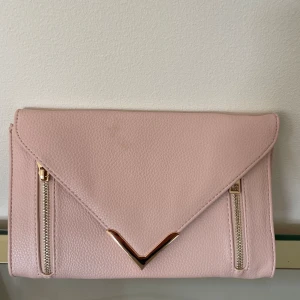 Ljusrosa kuvertväska  - Säljer en superfin ljusrosa kuvertväska. Väskan är gjord av syntetmaterial och har en elegant design med två dragkedjor på framsidan och en gulddetalj i mitten. Insidan är fodrad och har en liten ficka med dragkedja. Perfekt för fest eller andra speciella tillfällen! 💖