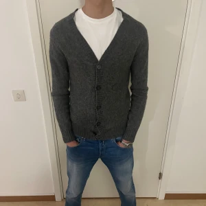 Grå kofta från Identity - Säljer en snygg och bekväm grå kofta från Identity. Den är gjord av 70% lammull och 30% polyamid, vilket gör den både varm och mjuk. Koftan har långa ärmar och knappar framtill. Perfekt för höst och vinter! 🧥