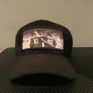 Svart keps med fototryck - Säljer en svart keps med ett coolt fototryck på framsidan. Kepsen har ett nätmaterial på baksidan för bättre ventilation och justerbar rem för perfekt passform. Märket 'AMORE' syns på insidan av kepsen. Perfekt för både vardag och sportaktiviteter.