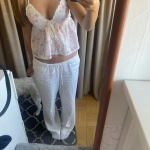 Söt blommig topp med spetsdetaljer - Säljer en söt blommig topp med spetsdetaljer. Toppen är vit med små rosa blommor och har en knytning framtill. Den är ärmlös och har en öppen rygg med korsade band. Perfekt för sommaren!
