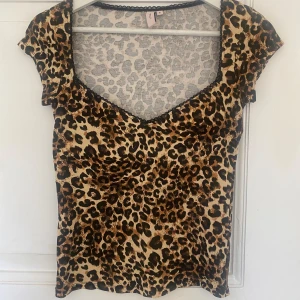 Leopardmönstrad topp från Nelly - Säljer en snygg leopardmönstrad topp från Nelly i storlek S. Toppen har en v-ringad hals och korta ärmar. Den är gjord av ett mjukt och bekvämt material, perfekt för en utekväll eller fest. Toppen har även en fin spetsdetalj vid halsringningen.