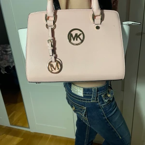 Rosa handväska från Michael Kors - Säljer en snygg rosa handväska den e inte äkta Michael Kors. Väskan har en stilren design med ett stort MK-logotyp i guld på framsidan. Den har två handtag och en justerbar axelrem. Perfekt för både vardag och fest! 💖