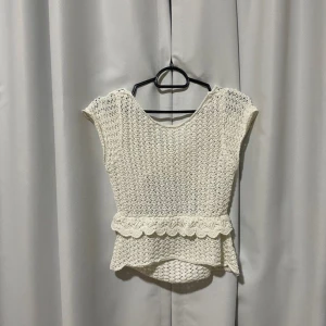 Vit stickad topp med volangdetaljer - Säljer en superfin vit stickad topp med volangdetaljer. Toppen är ärmlös och har en söt knytning framtill. Perfekt för sommaren och kan stylas både till vardags och fest. Den är i nyskick och väldigt bekväm att ha på sig. Passar perfekt till jeans eller kjol!