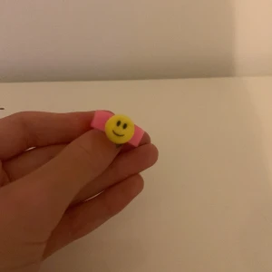 Rosa ring med smiley - Säljer en söt rosa ring med en gul smiley på. Perfekt för att lägga till en rolig och färgglad touch till din outfit. Ringen är gjord av plast och är i nyskick. Passar bra till både vardag och fest! är ganska liten