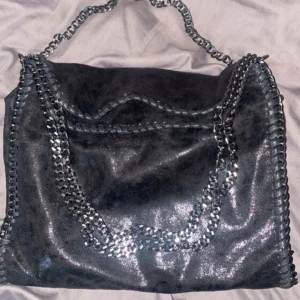 Svart glittrig axelväska med kedjedetaljer - Snygg svart axelväska i glittrigt material med coola kedjedetaljer. Perfekt för en utekväll eller fest! Väskan har en unik design med kedjor som handtag och längs kanterna, vilket ger den en edgy look. Den är rymlig och passar till alla dina essentials. Lik den trendiga Stockholmstil väskan 💕köptes för 700