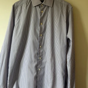 Randig skjorta från Eton - Säljer en stilren och klassisk randig skjorta från Eton i slim fit. Skjortan är blå och vit med långa ärmar och knappar framtill. Perfekt för både formella och avslappnade tillfällen. Skjortan är i mycket bra skick och gjord av högkvalitativt material.