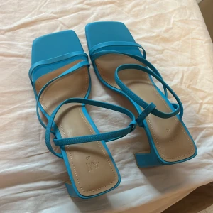 Blå sandaler med klack - Snygga blå sandaler med fyrkantig tå och smala remmar som korsar över foten. De har en elegant klack som ger lite extra höjd. Perfekta för sommarens fester eller en dag på stan. Skorna är i mycket bra skick och redo att användas!