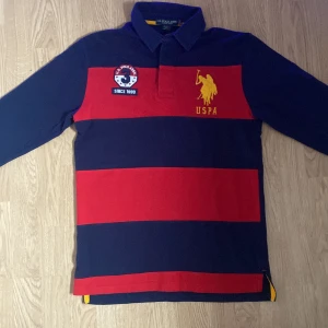 Polo pikétröja från U.S. Polo Assn. - Säljer en fet pikétröja från U.S. Polo Assn. i marinblått och rött. Tröjan har långa ärmar och ett stort gult tryck med USPA och siffran 3 på ryggen. I strl L men mäter S-M Hör av dig om du undrar om nått 🙌