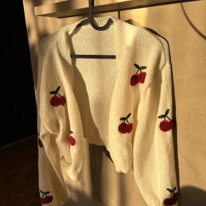 Vit kofta med körsbärsmönster - Superfin vit kofta med söta röda körsbär broderade över hela. Perfekt för att liva upp vilken outfit som helst! Den är stickad och har en mysig känsla, perfekt för kyliga dagar. Koftan har långa ärmar och en öppen design utan knappar. 