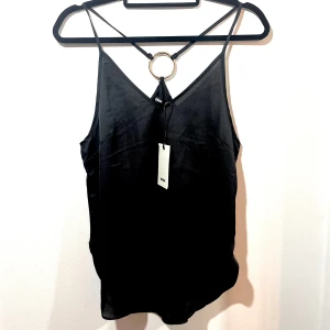 Svart linne med ringdetalj i guld. - Snyggt svart linne från Bikbok med en cool ringdetalj i ryggen. Perfekt för en utekväll eller fest! Linnet har tunna axelband och är i ett mjukt, lätt material som ger en skön känsla. 