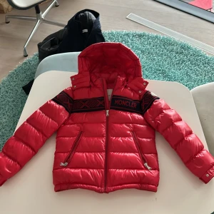 Röd dunjacka från Moncler - Säljer en snygg röd dunjacka från Moncler. Jackan har en cool design med blåa detaljer och Moncler-logga på ärmen. Den är perfekt för kalla dagar med sin varma vaddering och har en praktisk dragkedja framtill. Huvan är avtagbar och ger extra skydd mot väder och vind. Passar perfekt för vintern! Ny pris 17000 det låter dyrt men det finns inte många i världen!