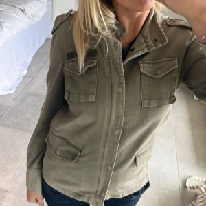 Olivgrön jacka från Anine Bing - Säljer en snygg olivgrön jacka från Anine Bing. Den har en cool militärstil med fyra fickor framtill och axelklaffar. Jackan är i bra skick och perfekt för höst och vår. Den har en dragkedja och knappar framtill för en stilren look. Passar perfekt till både vardag och fest!