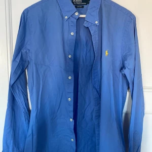 Blå skjorta från Polo Ralph Lauren - Säljer en snygg blå skjorta från Polo Ralph Lauren i slim fit. Skjortan har långa ärmar och klassiska knappar framtill. Den är i bomull och har en broderad gul logga på bröstet. Perfekt för både vardag och fest! 💙