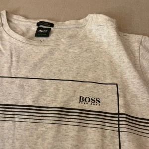 Grå t-shirt från Hugo Boss - Säljer en stilren grå t-shirt från Hugo Boss i mycket bra skick. Den har ett enkelt svart linjemönster över bröstet och korta ärmar. Perfekt för en avslappnad stil eller vardagsbruk.