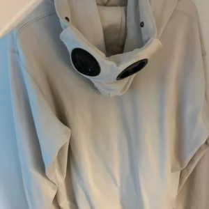 Beige hoodie från C.P. Company - Säljer en snygg beige hoodie från C.P. Company. Den har en unik design med glasögon i huvan, perfekt för en cool och avslappnad stil. Tröjan är i bra skick och passar perfekt för höst och vår. Den är långärmad och gjord i ett mjukt material som känns skönt mot huden.