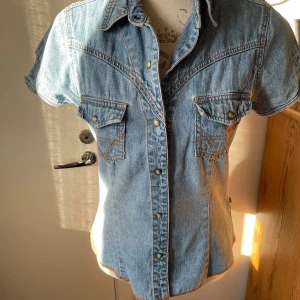 Jeanstopp från Clara - Snygg jeanstopp från Clara i 100% bomull. Den har korta ärmar och knappar framtill. Perfekt för en avslappnad stil med en touch av cowboy-vibe. Skjortan har två bröstfickor med knappdetaljer och en klassisk krage. Perfekt för vår och sommar!