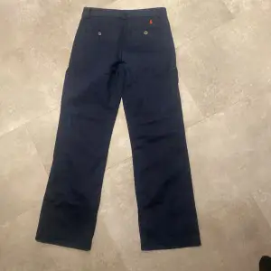 Säljer ett par snygga mörkblå byxor från Polo Ralph Lauren. De har en klassisk straight passform och är tillverkade i bomull. Byxorna har knappar och en broderad röd logga på baksidan. Perfekta för både vardag och fest!