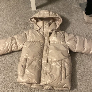 Beige pufferjacka från 157 - Säljer en snygg beige pufferjacka från 157 i storlek 150. Jackan har en glansig yta och är perfekt för kalla dagar. Den har en huva och långa ärmar, samt praktiska fickor framtill. Perfekt för vintern! ❄️ passar en xs eller s☺️❤️