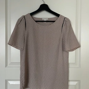 Beige blus med mönster - Säljer en söt beige blus med små svarta mönster. Blusen har korta ärmar och en rund halsringning. Perfekt för både vardag och festliga tillfällen. Den är i mycket bra skick och passar perfekt till både jeans och kjol.