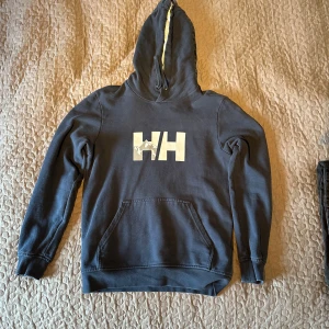 Mörkblå hoodie från Helly Hansen - Säljer en snygg mörkblå hoodie från Helly Hansen i bra skick. Den har en stor logga på framsidan och en praktisk magficka. Perfekt för kyliga dagar eller som en casual vardagströja. Huvan är fodrad för extra värme.