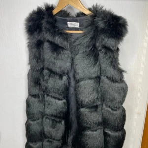 Svart fusk pälsväst - Säljer en supermjuk och fluffig svart pälsväst som är perfekt för kyliga dagar. Västen har en lyxig känsla och är i mycket bra skick. Den är utan knappar eller dragkedja, vilket ger en öppen och avslappnad stil. Passar perfekt över en tröja eller klänning för en chic look.