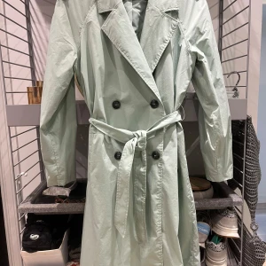 Ljusgrön trenchcoat från Pimkie - Säljer en superfin ljusgrön trenchcoat från Pimkie. Den har en klassisk design med knappar och ett bälte i midjan för en snygg passform. Perfekt för vår och höst! 💚