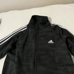 Svart träningsjacka från Adidas - Säljer en svart träningsjacka från Adidas i mycket bra skick. Den har de klassiska tre vita ränderna längs ärmarna och en dragkedja framtill. Perfekt för träning eller som en casual jacka. Adidas-loggan är broderad på bröstet. Passar bra till både höst och vår.