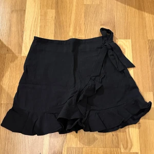 Svart omlottkjol med volang - Säljer en superfin svart omlottkjol med volangdetaljer. Perfekt för både vardag och fest! Kjolen har en knytning i sidan som ger en snygg passform. Den är kort och passar perfekt till sommaren eller en utekväll. 🖤
