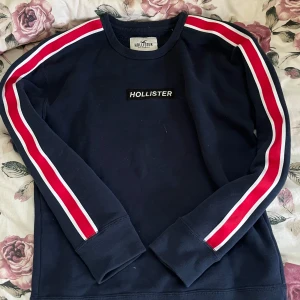 Mörkblå tröja från Hollister - Hollister tröja med röda och vita ränder längs ärmarna. 