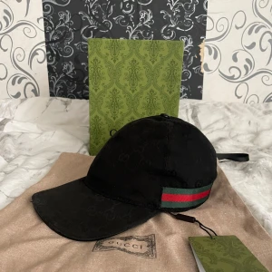 Svart keps från Gucci - Snygg svart keps från Gucci med deras ikoniska mönster. Den har en justerbar rem baktill och en grön och röd rand på sidan. Perfekt för att ge din outfit en lyxig touch!