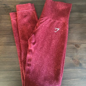 Röda leggings från Gymshark - Säljer ett par snygga röda leggings från Gymshark i storlek XS. De är perfekta för träning med sin tighta passform och bekväma midja. Materialet är mjukt och stretchigt, vilket gör dem ideala för alla typer av aktiviteter. De har en diskret logga på benet. Perfekta för gymmet eller yoga!